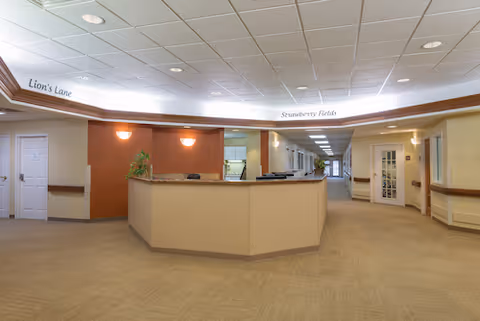 Reception desk in a senior living facility foyer with a long corridor and overhead signs reading 'Lion's Lane' and 'Strawberry Fields'.