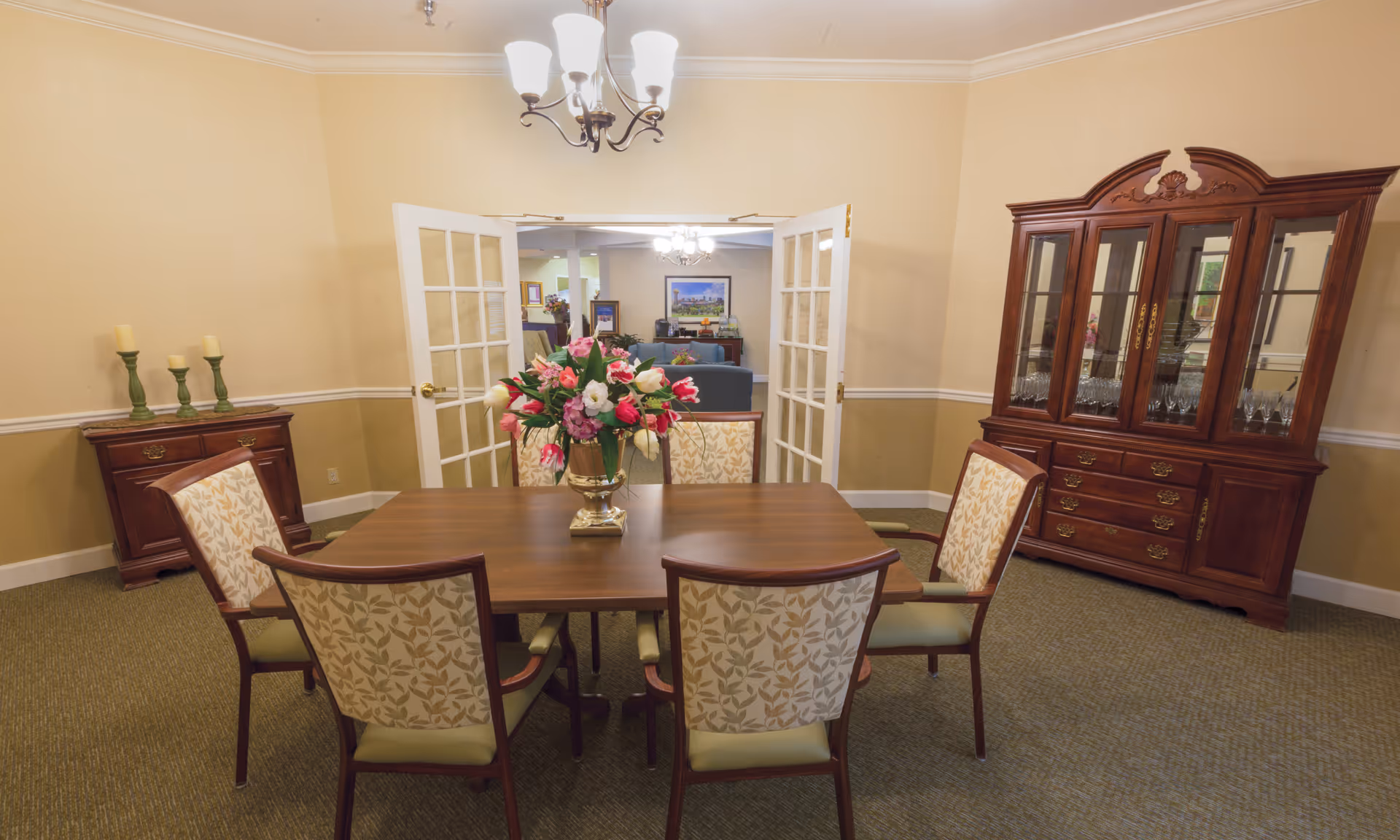 A dining room with a wooden table surrounded by six upholstered chairs with leaf-patterned fabric. A vase with a colorful bouquet of flowers is placed in the center of the table. The room has beige walls with white trim, a chandelier hanging from the ceiling, a wooden cabinet with candles on the left, and a large wooden china cabinet with glass doors on the right. French doors open to a living area with sofas and a framed picture on the wall.