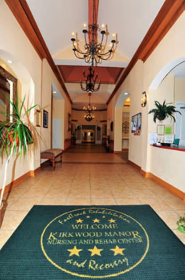 Interior view of the entrance hallway of Kirkwood Manor Nursing and Rehab Center featuring a green welcome mat with the facility's name, beige walls, tiled floor, chandeliers hanging from the ceiling, a bench along the left wall, and a reception window on the right with a plant nearby.