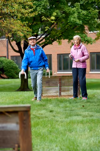 Two elderly individuals playing a lawn game on a grassy area outside, with trees and a brick building in the background. One person is wearing a blue jacket and gray pants, and the other is wearing a pink jacket and dark pants.