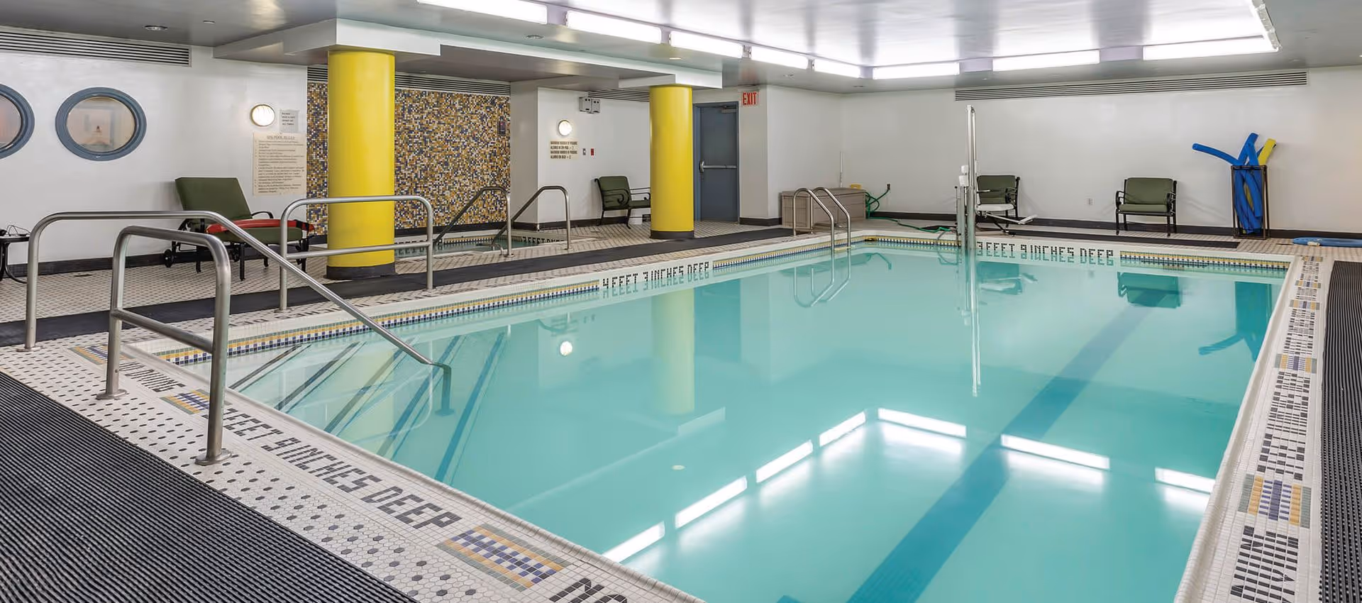 Indoor swimming pool area with clear blue water, metal handrails, yellow columns, mosaic tile accents, and several green lounge chairs along the walls.