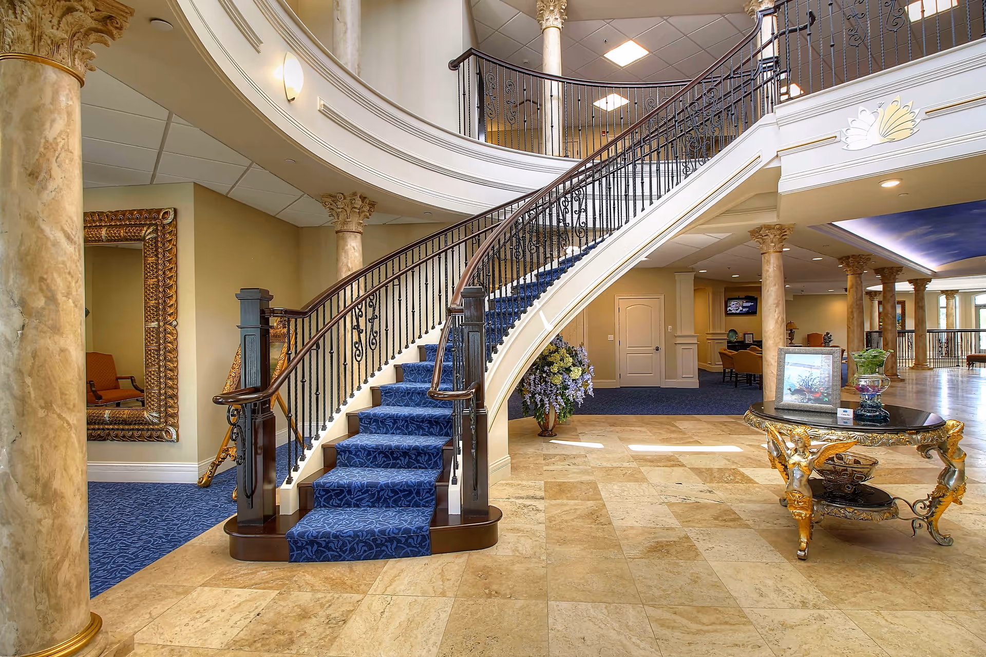 Elegant interior of Tranquility Estates featuring a grand curved staircase with blue carpet, ornate gold-trimmed table with decorative items, marble columns, and a large mirror on the wall. The space has a polished tile floor and a blue carpeted area in the background with seating and a television.