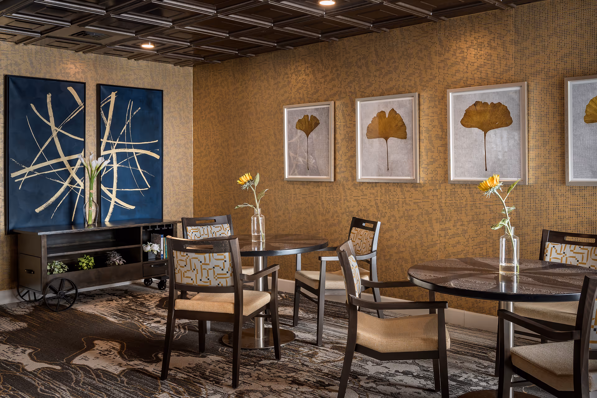 A cozy dining area with round dark wood tables and cushioned chairs featuring patterned upholstery. Each table has a single vase with a sunflower. The walls are decorated with framed botanical prints of leaves and two large abstract blue and beige paintings. The floor has a patterned carpet and there is a dark wood sideboard with plants and books underneath the paintings.