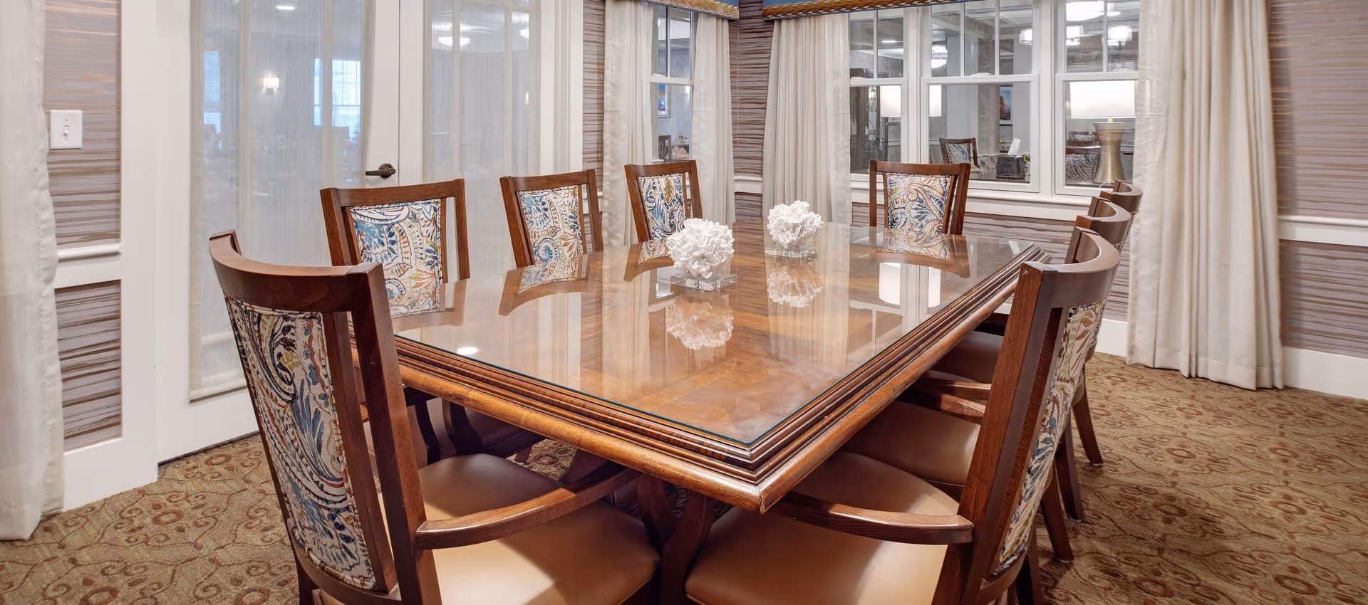 A polished wooden dining table surrounded by upholstered chairs in an elegant dining room with patterned carpet and curtained windows.