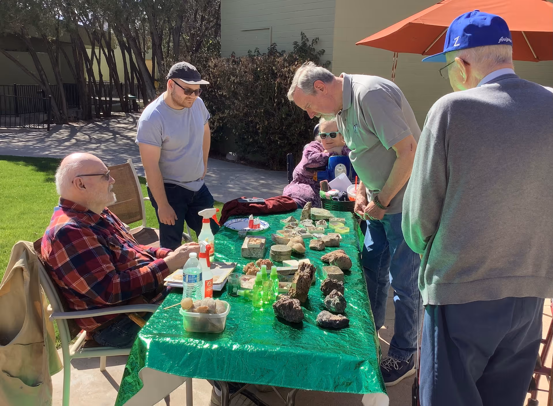 A group of five people gathered around a table covered with a green tablecloth outdoors. The table displays various rocks and minerals. One man in a red plaid shirt is seated, while others stand around the table, engaging and observing the items. The setting is sunny with trees and a building in the background.