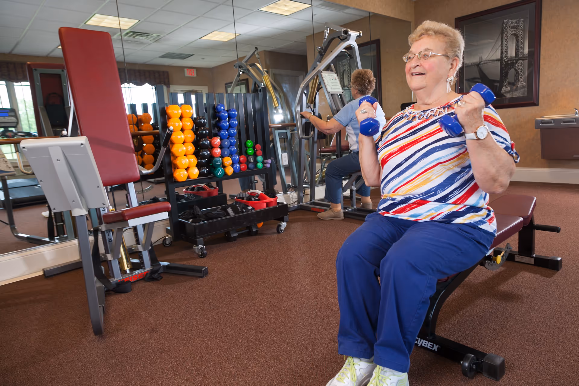 An elderly woman wearing a colorful striped shirt and blue pants is sitting on a workout bench in a fitness room, lifting blue dumbbells. Behind her, there is a rack with various colorful dumbbells and exercise equipment, with mirrors on the wall reflecting the scene.