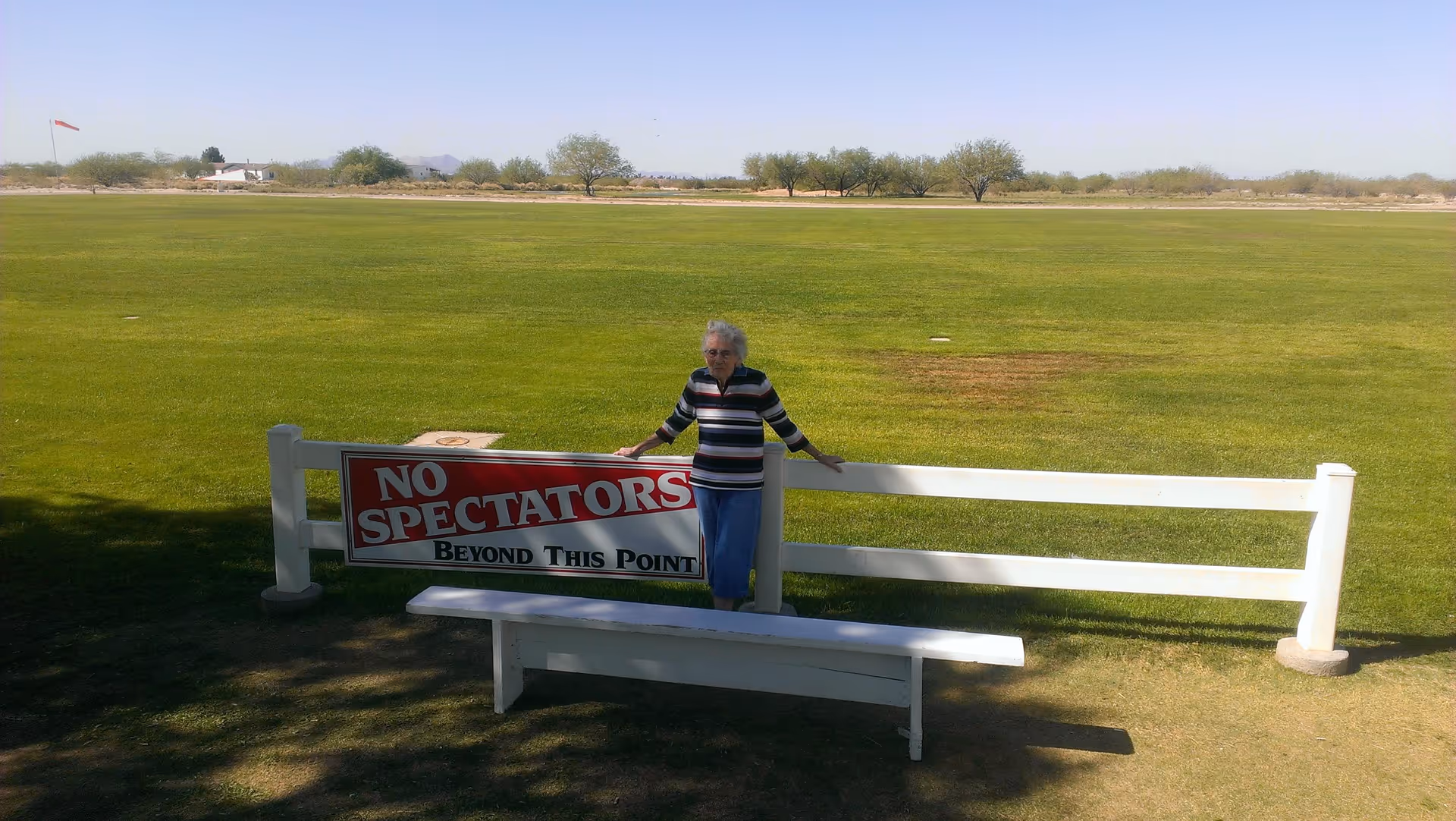 An elderly woman standing next to a white fence with a sign that reads 'NO SPECTATORS BEYOND THIS POINT' in front of a large green field with trees and a clear sky in the background.