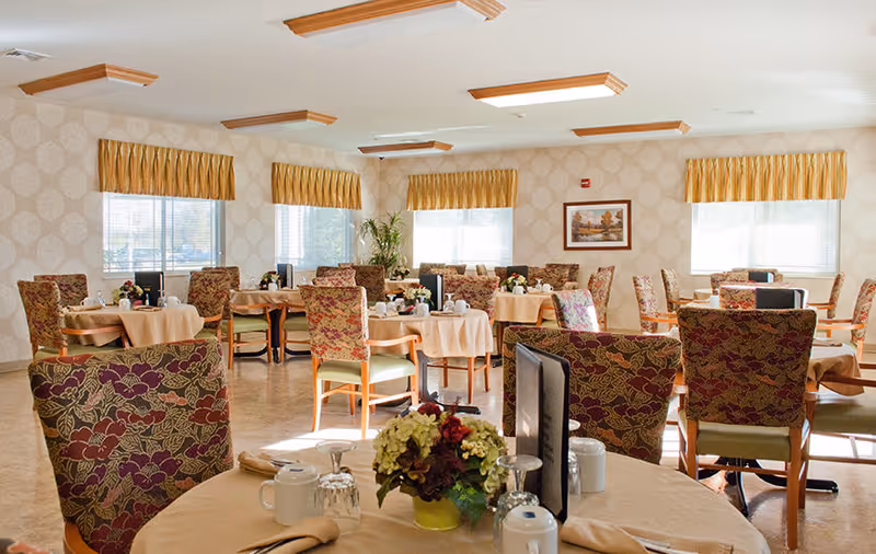 A bright dining room with multiple round tables covered with beige tablecloths, each set with cups, glasses, and napkins. The chairs have floral patterned upholstery. The room has large windows with yellow valances, patterned wallpaper, and a framed picture on the wall. A floral centerpiece is visible on the nearest table.