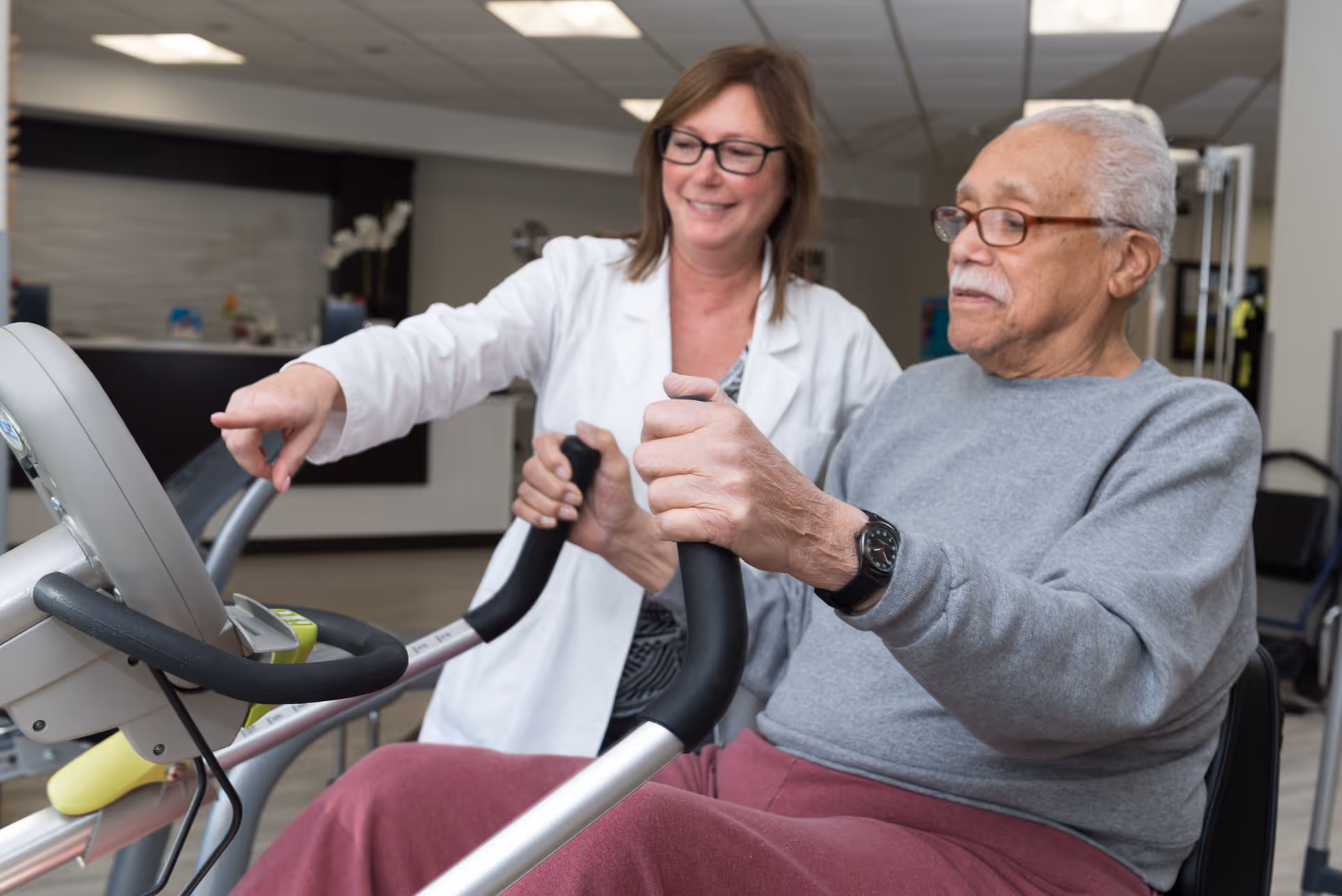 An elderly man wearing glasses and a gray sweater is using a stationary exercise bike while a female healthcare professional in a white coat smiles and points at the bike's display screen, assisting him in a rehabilitation or nursing center setting.