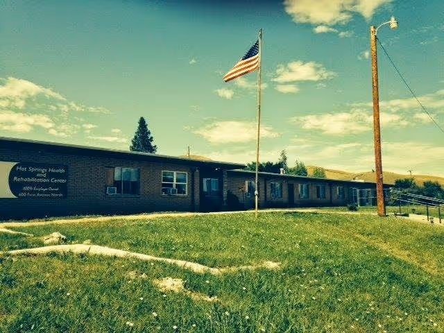Exterior view of Hot Springs Health and Rehabilitation Center, a single-story building with multiple windows, an American flag on a flagpole in front, and a grassy area with a sidewalk leading to the entrance under a partly cloudy sky.