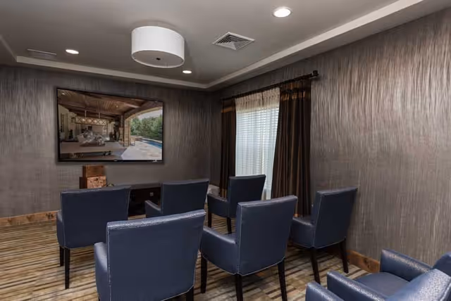 A small media room with six blue upholstered chairs arranged in two rows facing a wall-mounted flat screen TV. The room has textured gray walls, a window with brown and beige curtains, and a modern ceiling light fixture. The floor has a striped carpet with shades of beige and brown.