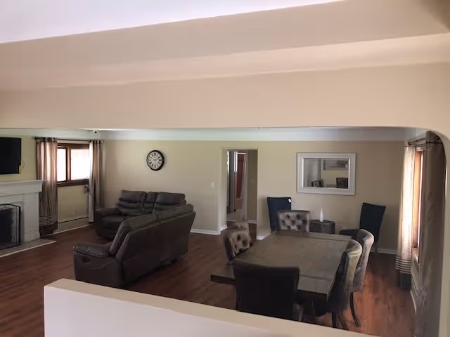 A spacious living room area with wooden flooring featuring a dark brown sofa set near a fireplace on the left side. A dining table with six upholstered chairs is positioned on the right side of the room. The walls are painted beige, with a clock hanging on the far wall and a large mirror on the right wall. Windows with curtains allow natural light into the room.