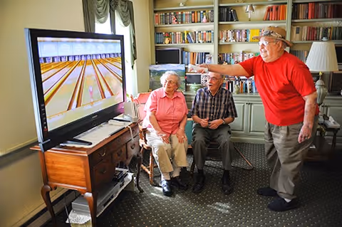 Three elderly people in a living room playing a bowling video game on a large TV. Two are seated on chairs, watching, while one stands and points at the screen. The room has bookshelves filled with books and a window with curtains.