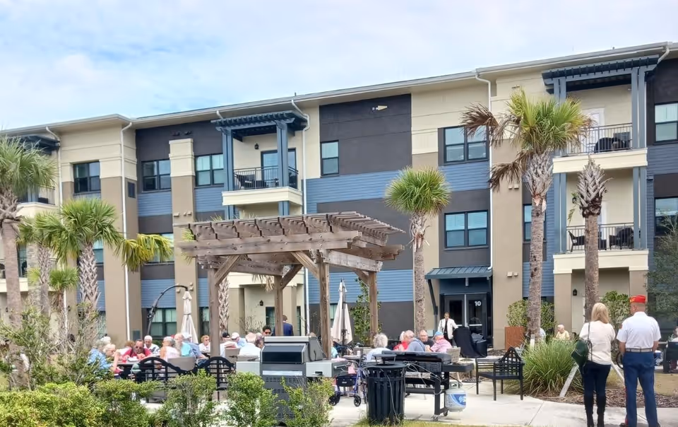 Outdoor patio area at Oak Hill Senior Living with a group of seniors sitting and socializing under a wooden pergola. The background shows a three-story residential building with balconies and palm trees surrounding the area.