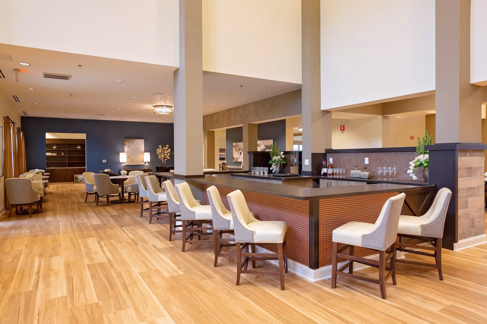A spacious and well-lit common area in Jubilee House featuring a long bar counter with cushioned bar stools, a wooden floor, and a seating area with armchairs and tables in the background. The walls are painted in neutral tones with decorative artwork and flowers placed on surfaces.
