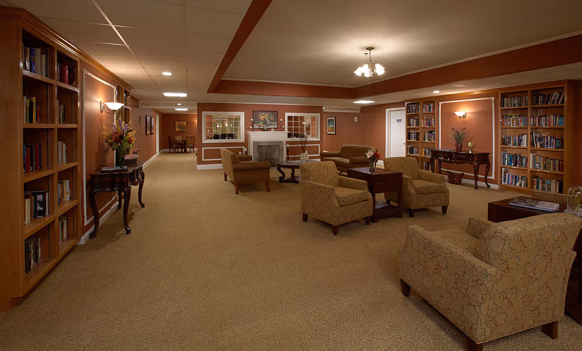A cozy living room area in a senior living facility with beige carpet, patterned armchairs, sofas, wooden coffee tables, and built-in wooden bookshelves filled with books. The room features warm brown walls, a white fireplace with a painting above it, and soft lighting from ceiling fixtures and wall sconces.