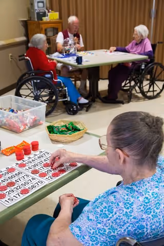 Four elderly individuals, three in wheelchairs, are seated around two tables playing bingo. The foreground shows a woman in a blue patterned shirt marking her bingo card with red chips. The background shows two women and one man engaged in the game, with a container of bingo chips and a basket of snacks on the tables. The setting appears to be a communal activity room with beige walls and a curtain in the background.