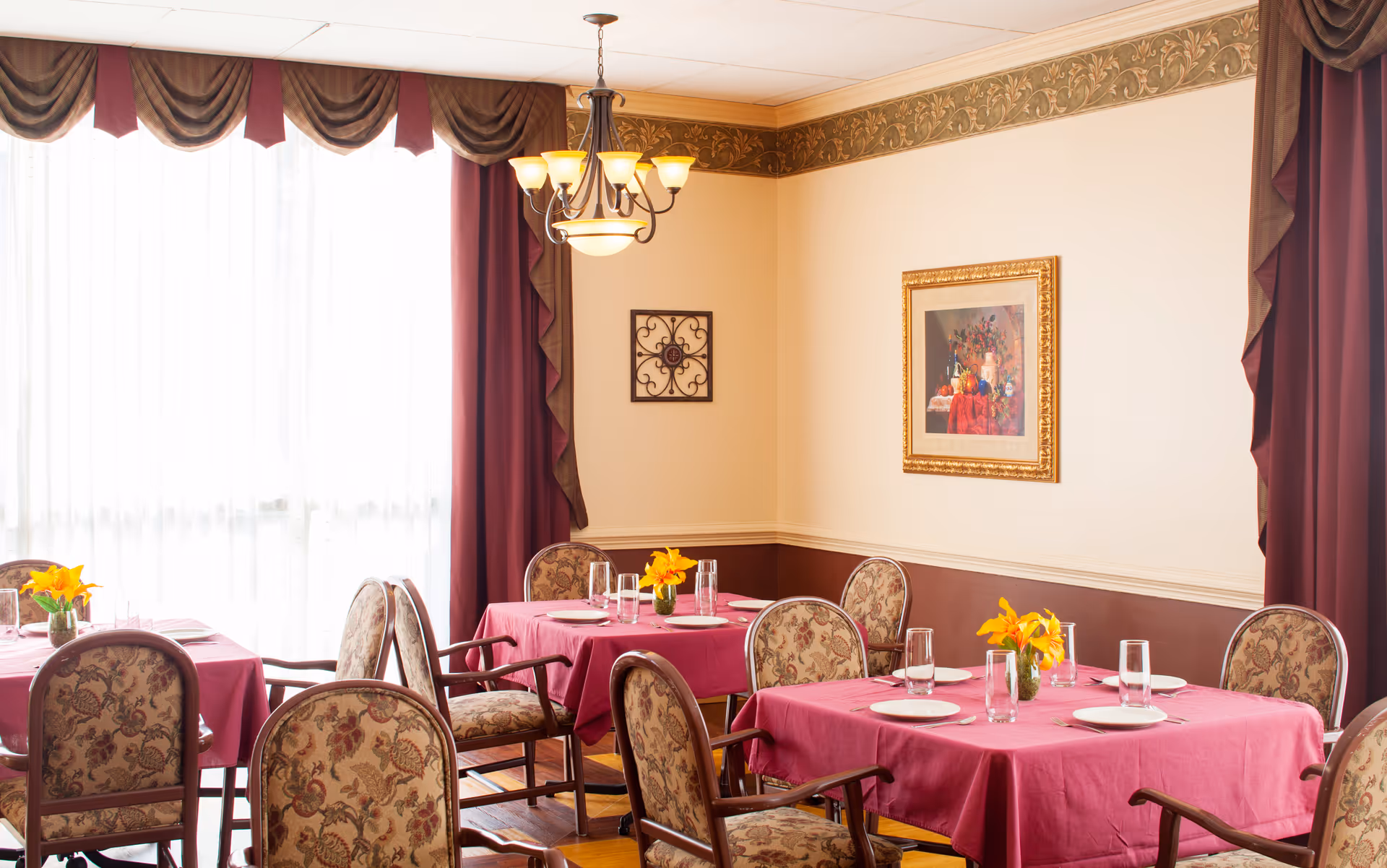A dining room with several tables covered in pink tablecloths, each set with plates, glasses, and small vases of yellow flowers. The room has floral upholstered chairs, large windows with sheer white curtains and burgundy drapes, a chandelier hanging from the ceiling, and framed artwork on the cream-colored walls.