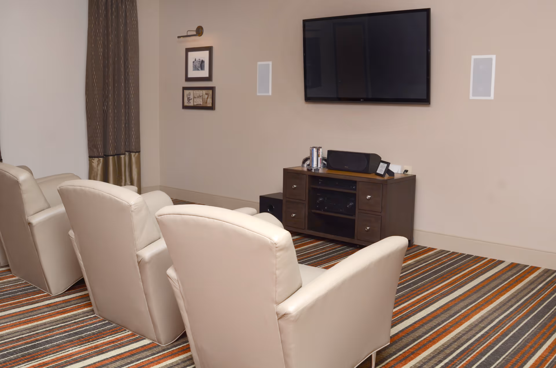 A cozy room with three beige recliner chairs facing a wall-mounted flat screen TV. Below the TV is a wooden cabinet with electronic devices and speakers. The room has a striped carpet in shades of brown, orange, and gray, and two framed pictures on the wall next to a window with brown curtains.
