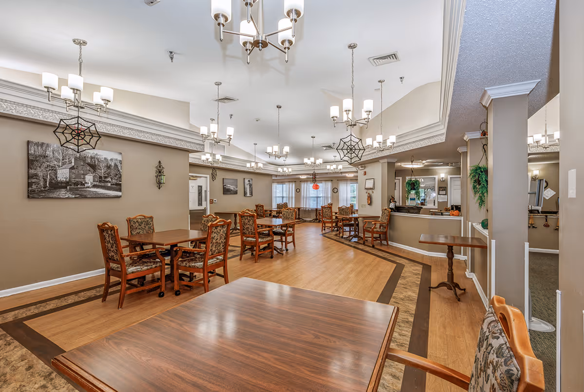 A spacious dining room with multiple wooden tables and upholstered chairs arranged neatly. The room features beige walls, decorative wall art, hanging chandeliers with multiple lights, and some Halloween-themed decorations like spider webs and pumpkins. There is a reception or service counter on the right side with plants and additional decor.
