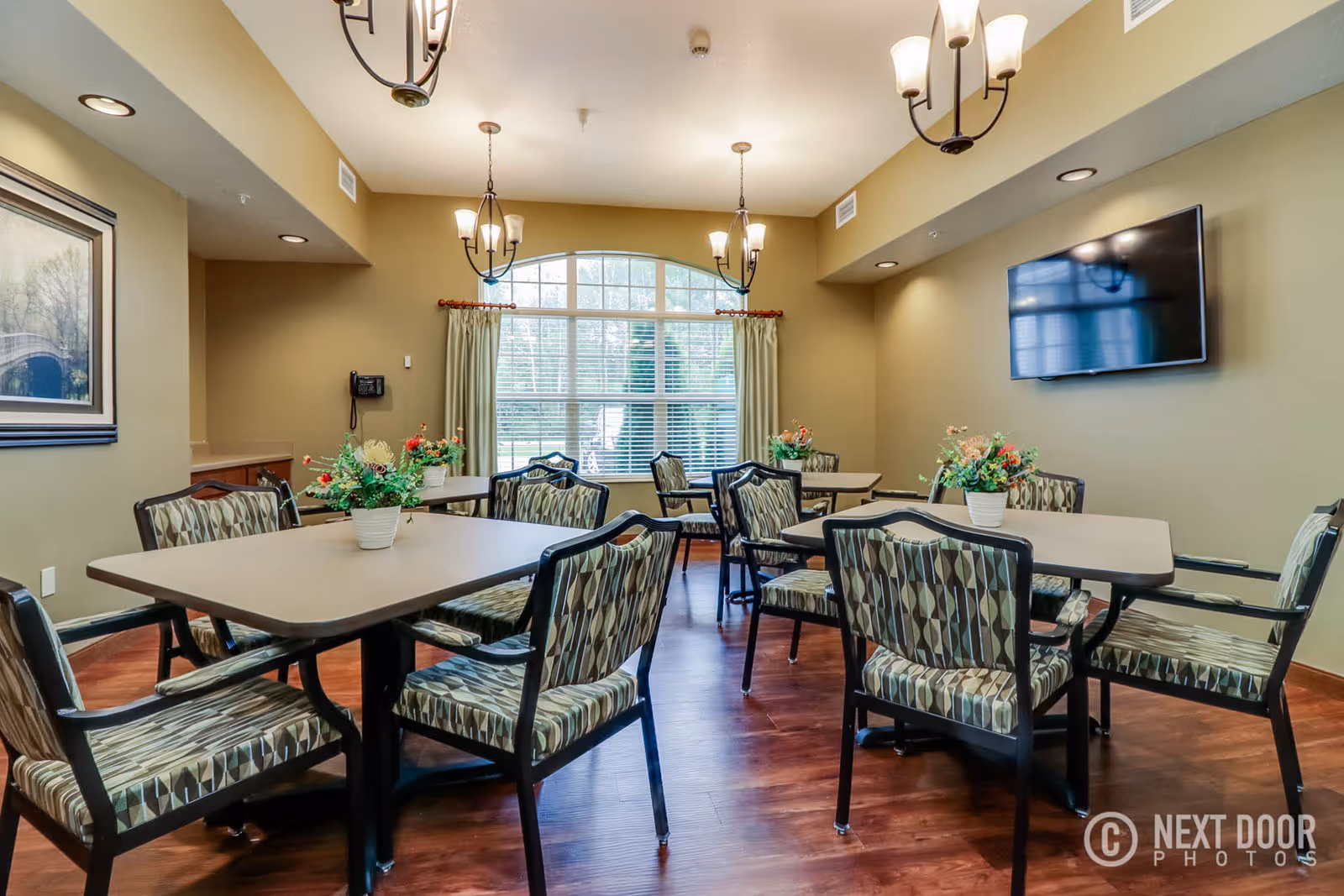 Sunlit communal dining/activity room with multiple tables and patterned chairs, potted flowers, chandeliers, and a wall-mounted TV.
