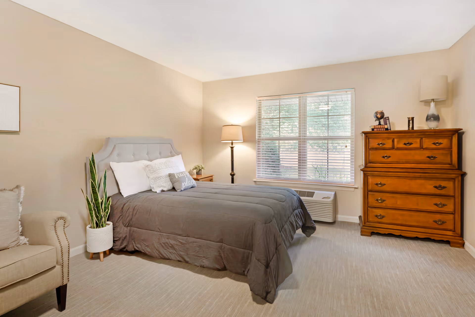 A cozy bedroom with a gray upholstered bed featuring white and gray pillows, a wooden dresser with decorative items on top, a floor lamp, a large window with blinds, a potted plant, and a beige armchair partially visible on the left.