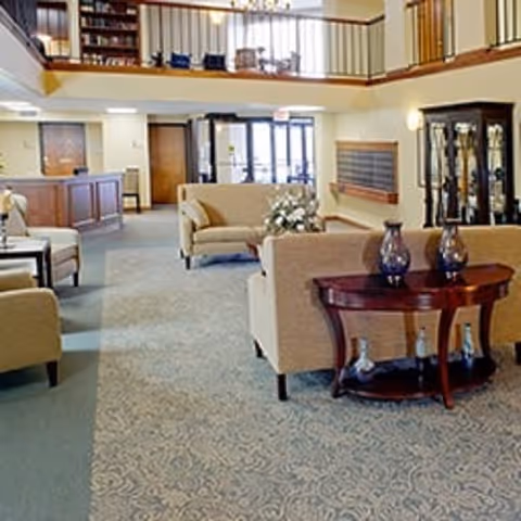 Spacious senior living facility common area with beige upholstered sofas and armchairs arranged around a wooden table with decorative vases. The room has a patterned carpet, a reception desk on the left, and a second-floor balcony with bookshelves and seating visible above.