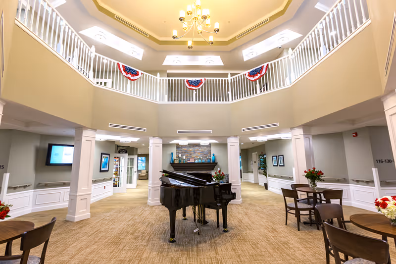 Spacious, well-lit common area in a senior living facility featuring a black grand piano in the center, round tables with chairs and flower arrangements, white railings on the upper level decorated with red, white, and blue bunting, beige walls, and a chandelier hanging from the ceiling.