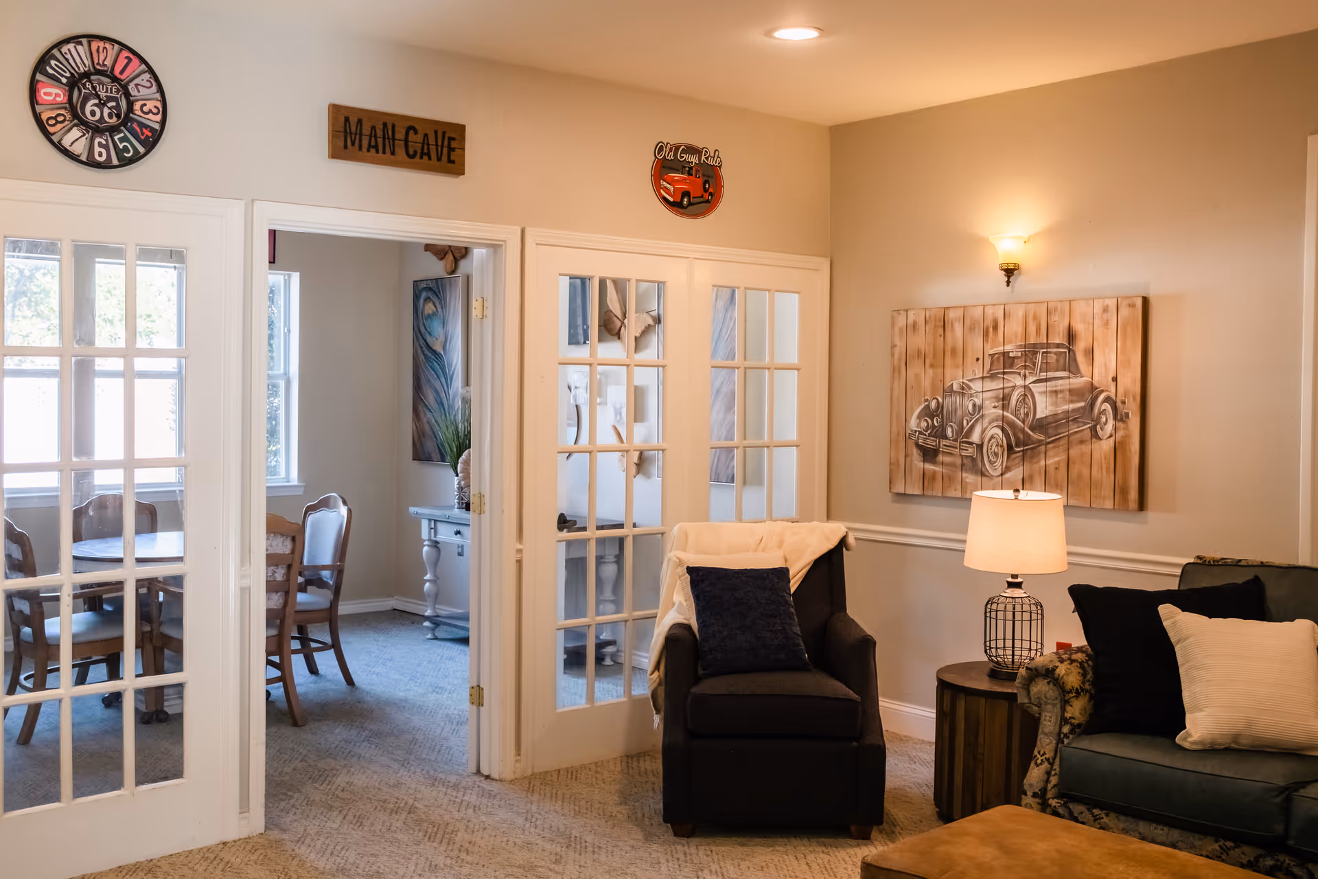 Interior view of a cozy living area with a dark armchair draped with a light blanket and a dark pillow, next to a round wooden side table with a lamp. A green patterned sofa with multiple pillows is partially visible. On the wall above the armchair is a wooden art piece depicting a vintage car. To the left, there are glass-paneled double doors leading to a dining room with a round wooden table and chairs. Above the doorways are decorative signs, including one that says 'MAN CAVE' and another with a vintage truck and the words 'Old Guys Rule'. A colorful Route 66 clock is mounted on the wall near the ceiling.