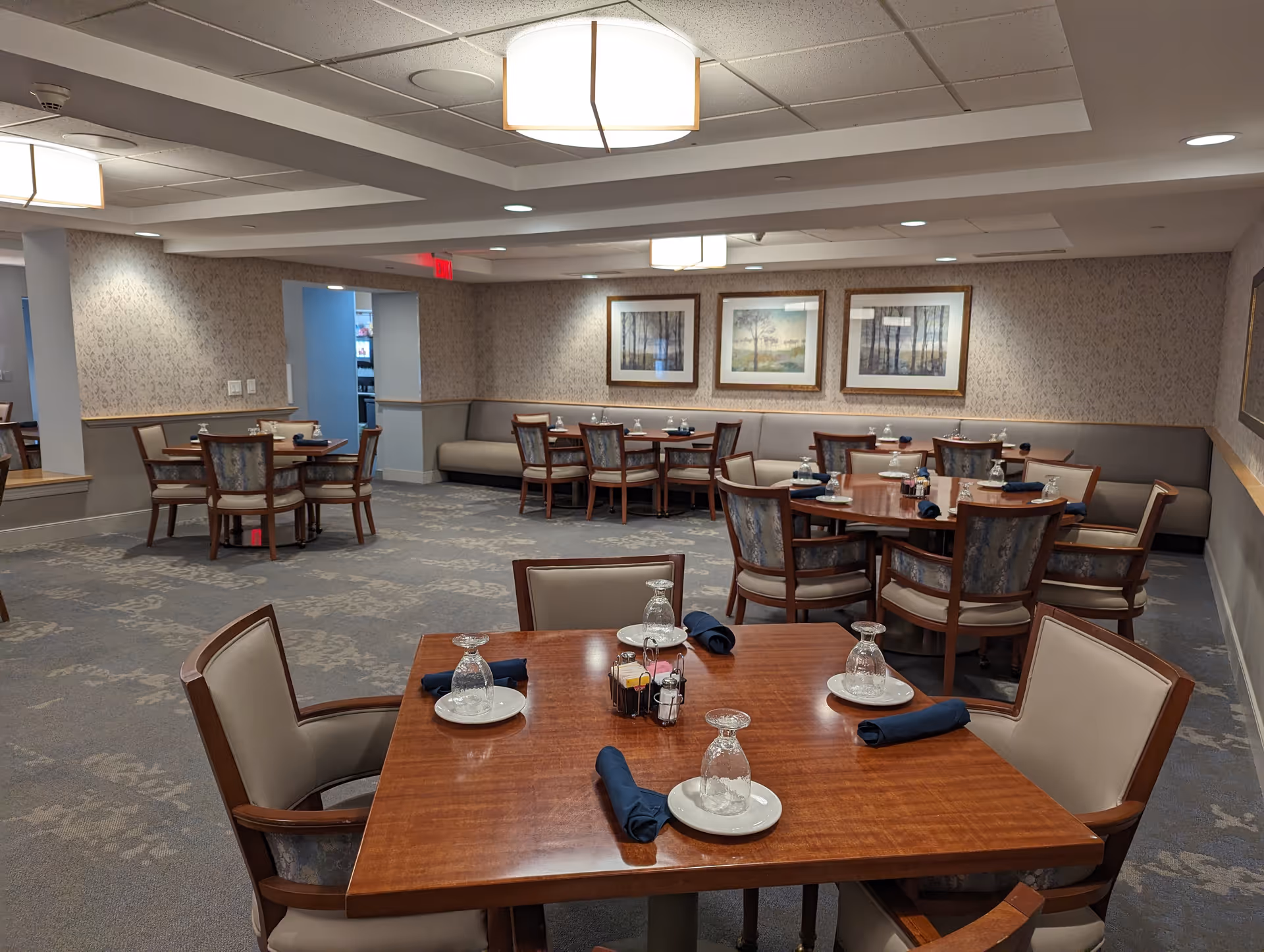 A dining room in a senior living facility with several wooden tables and cushioned chairs arranged neatly. Each table is set with upside-down glasses, white plates, and dark blue napkins. The room has patterned carpet, wallpapered walls with framed landscape artwork, and soft ceiling lighting.