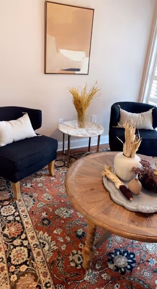 A cozy living room area with two dark blue armchairs each with a white pillow, a round wooden coffee table with decorative dried plants and corn, a small round side table with a vase of dried wheat, an abstract painting on the wall, and a patterned red and beige area rug.