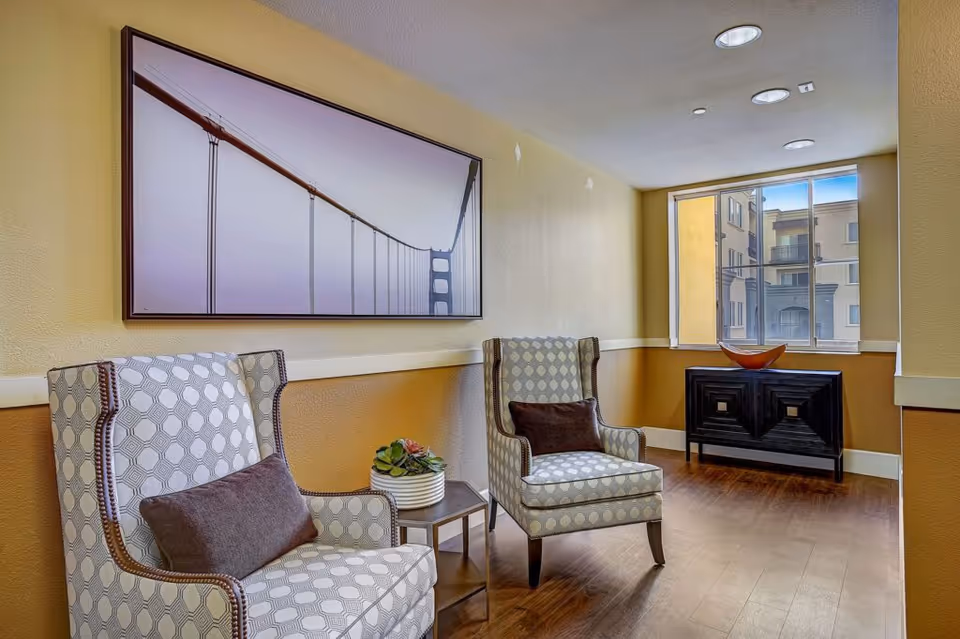 A cozy sitting area with two patterned armchairs each with a brown cushion, a small side table with a potted plant, a large framed picture of a suspension bridge on the wall, a window showing an exterior view of nearby buildings, and a dark wooden cabinet with a decorative bowl on top.