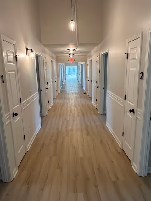 A long, well-lit hallway with light wood flooring and white walls. There are multiple white doors on both sides of the hallway, each door numbered. Modern light fixtures hang from the ceiling and are mounted on the walls. At the end of the hallway, there is a window letting in natural light.
