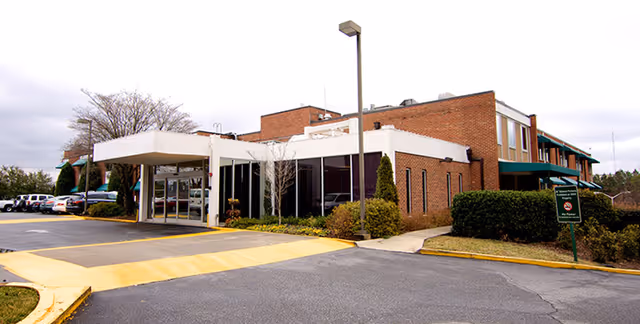 Exterior view of a brick building with a covered entrance and parking area, surrounded by bushes and trees under a cloudy sky.