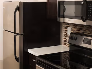 A kitchen corner showing a stainless steel refrigerator, microwave, electric stove, and mosaic tile backsplash.