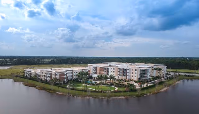 Aerial view of a large senior living facility named Watercrest Palm Beach Gardens, surrounded by water on three sides with landscaped green areas and palm trees, under a partly cloudy sky.