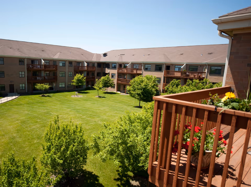 View of a large grassy courtyard surrounded by a three-story brick building with balconies. The foreground shows a wooden balcony with flower pots containing red and yellow flowers. Several small trees are scattered throughout the courtyard under a clear blue sky.