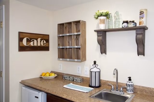 A small cafe-style countertop area with a sink, faucet, and soap dispenser. Above the counter is a wooden shelf holding decorative items including a potted plant, glass bottles, and jars. A wooden wall-mounted organizer with numbered cubbies is on the wall next to a wooden sign that says 'Cafe'. On the counter, there is a bowl of lemons, a decorative lantern, and a sign that reads 'Coffee and friends a perfect blend'.
