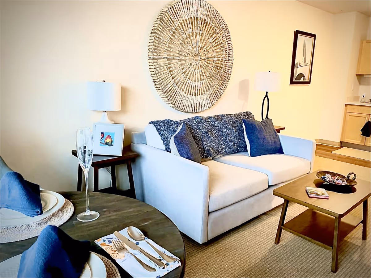 A cozy living room area featuring a light-colored sofa with blue cushions and a textured throw blanket. A wooden coffee table with decorative items sits in front of the sofa. To the left, a round dining table is set with plates, silverware, and a champagne flute. The room is decorated with a large circular wall art piece above the sofa, two table lamps on side tables, and a framed picture on the wall. The background shows part of a kitchen area with light wood cabinetry.