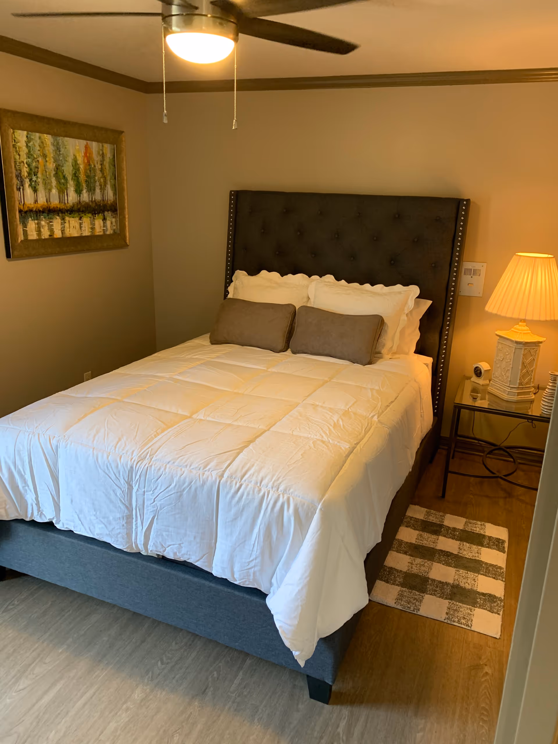 A cozy bedroom with a neatly made bed featuring a dark upholstered headboard, white bedding, and two gray pillows. A bedside table with a decorative lamp and small items sits to the right of the bed. A ceiling fan with a light is above, and a framed painting of trees hangs on the left wall. A small checkered rug is placed on the wooden floor beside the bed.