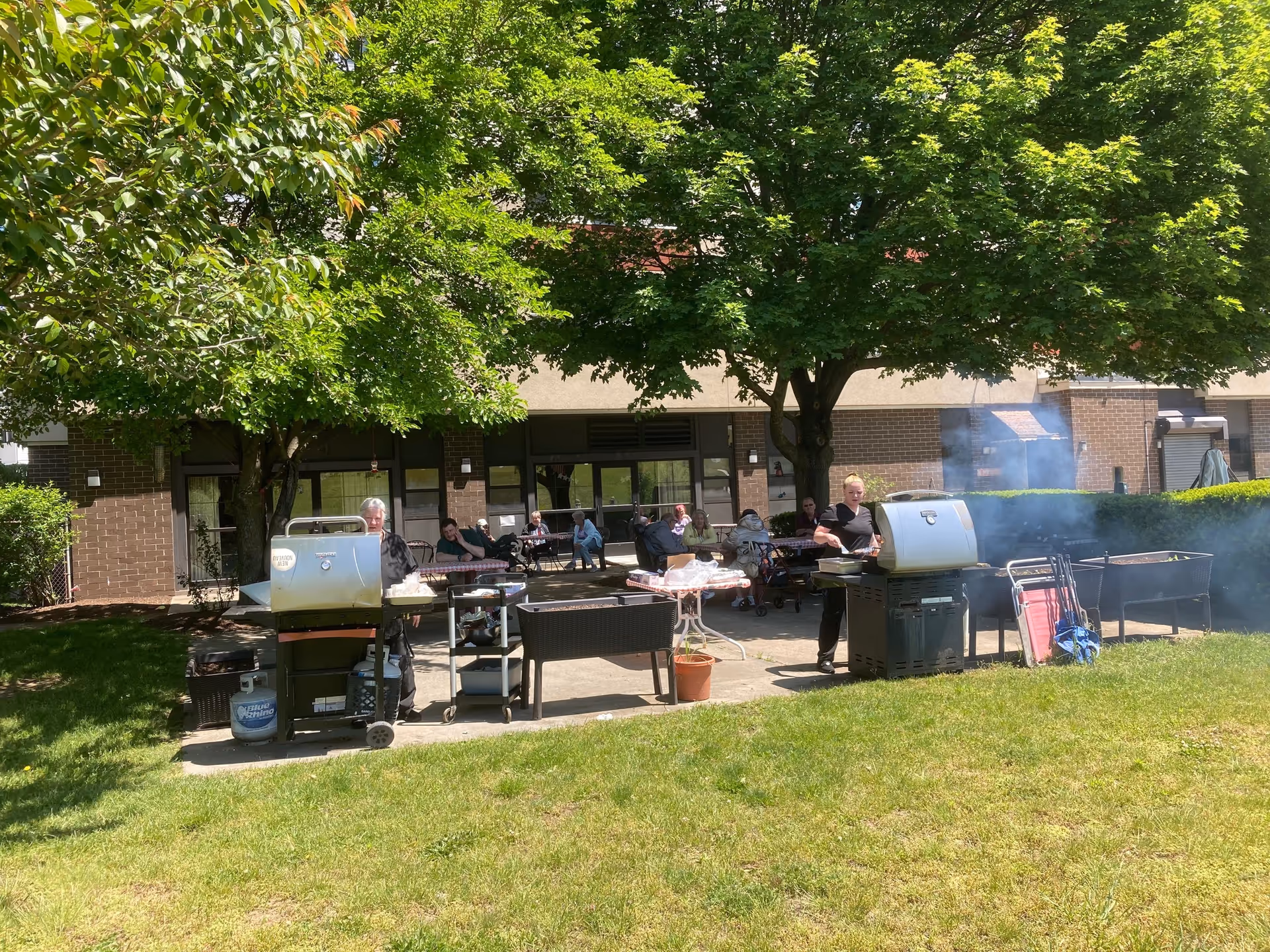 Outdoor patio area at St. Germain Manor with several people sitting at tables under large green trees. Two individuals are grilling food on gas grills with smoke rising. The building with large windows and brick walls is visible in the background.