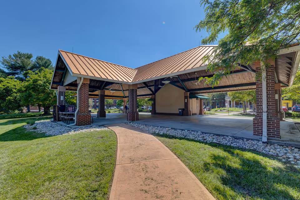 A covered outdoor pavilion with brick columns and a metal roof on landscaped grounds with a walkway leading to it.