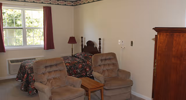 A bedroom with two beige cushioned armchairs and a small wooden table between them. In the background, there is a bed with a floral bedspread, a wooden headboard, a floor lamp with a red lampshade, a window with red curtains, and an air conditioning unit below the window. A wooden wardrobe is visible on the right side of the image.