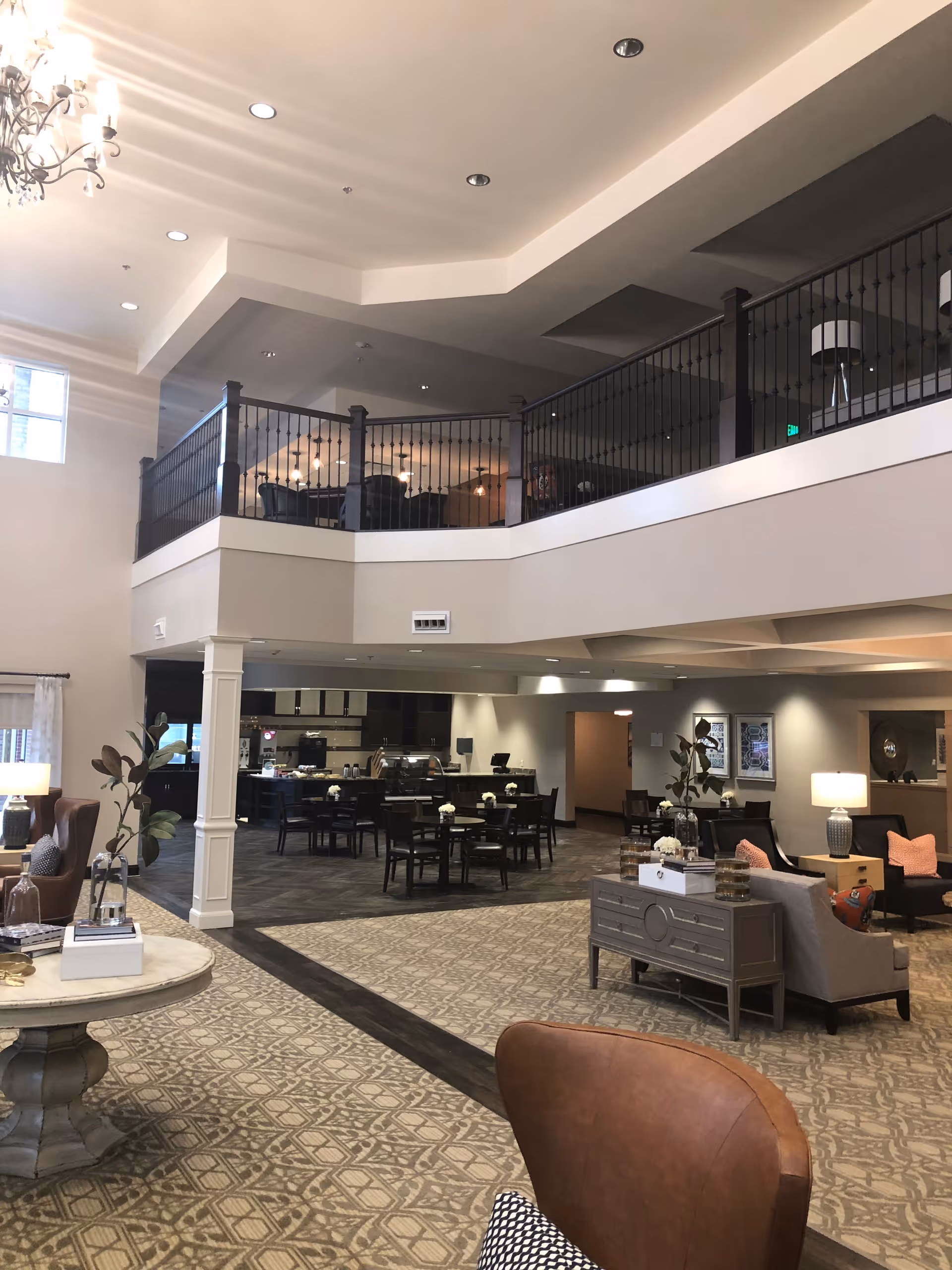 Spacious and well-lit interior of a senior living facility featuring a seating area with armchairs and sofas, a round table with decorative items, and a dining area with multiple tables and chairs. The space has a high ceiling with recessed lighting and a chandelier, and an upper balcony with black railings overlooking the area below.