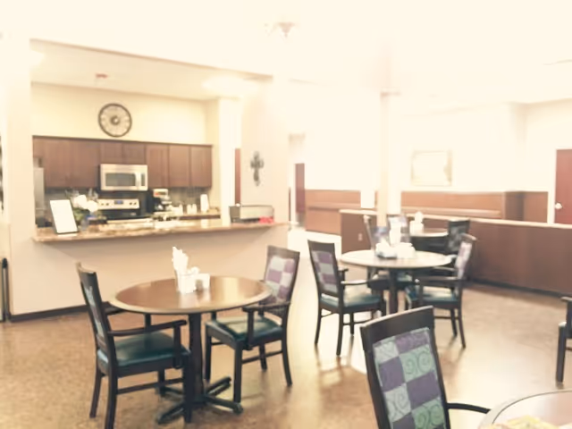 Interior view of a dining area with several round tables and chairs. The chairs have patterned upholstery in shades of purple and green. In the background, there is a kitchen area with wooden cabinets, a microwave, and a stove. The room is well-lit with natural light coming from windows.