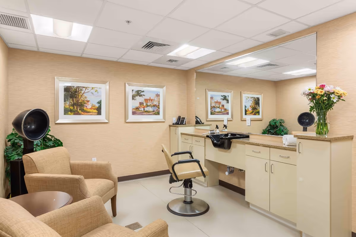 A well-lit hair salon room with beige walls and flooring. The room features a salon chair in front of a large mirror and a black hair washing sink. There are two beige armchairs and a small round table on the left side, along with a black hair dryer hood. The walls are decorated with framed landscape paintings, and a vase of colorful flowers sits on the countertop.