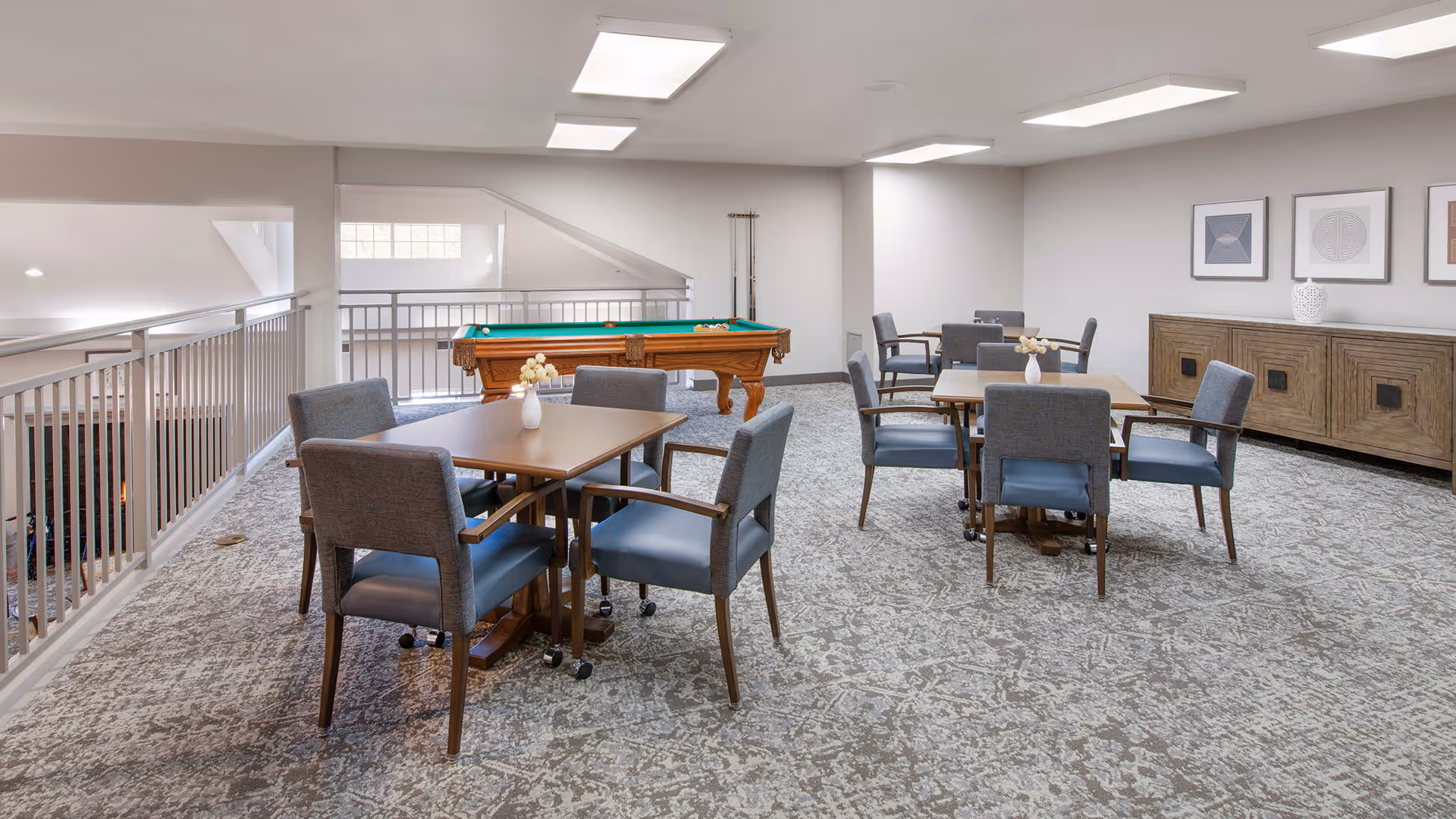 A spacious recreational room with carpeted floor featuring two square tables each surrounded by four gray cushioned chairs. A pool table with green felt is positioned near the railing overlooking a lower level. The walls are light-colored with three framed abstract artworks and a wooden sideboard with decorative items on top. The room is well-lit with ceiling lights.