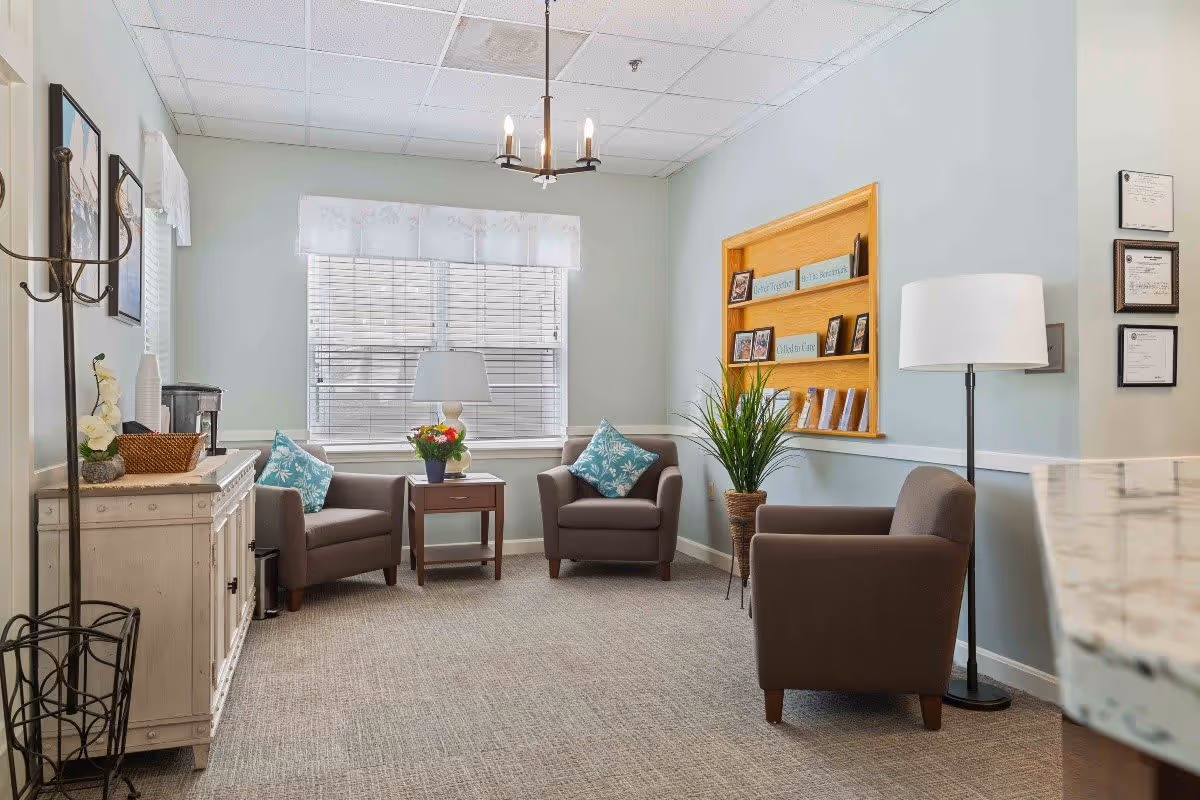 A cozy sitting area in a senior living facility with three brown armchairs, two with blue patterned pillows, arranged around a small wooden side table with a lamp and a flower pot. The room has light blue walls, a window with white blinds and a valance, a white sideboard with decorative items, a tall floor lamp, a plant in a woven basket, and framed certificates on the wall. A chandelier hangs from the ceiling.