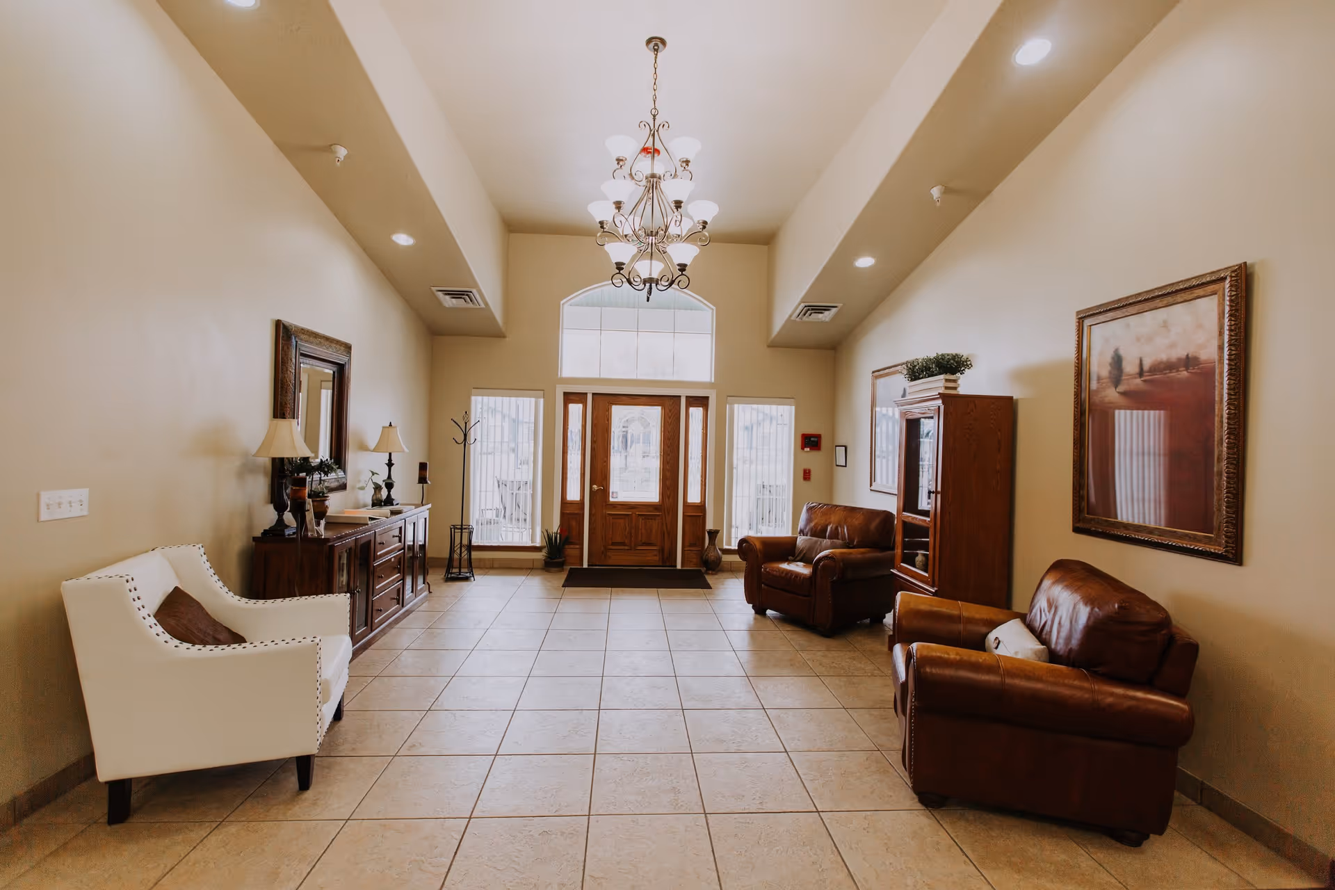 Bright tiled entry lobby with a chandelier, white and leather armchairs, wooden cabinets, and double wooden front doors.