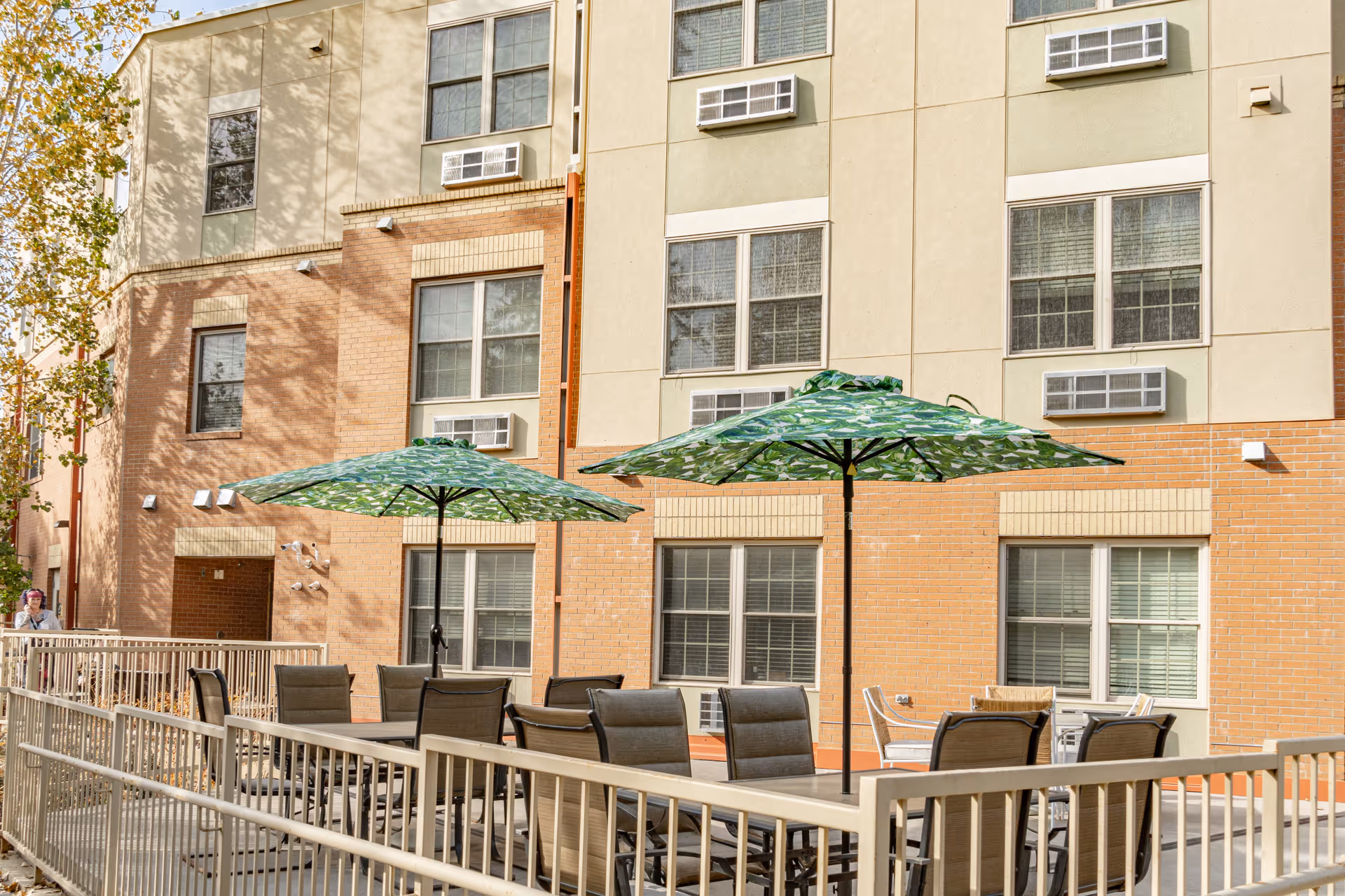 Outdoor patio area at Cedar Ridge Place with multiple tables and chairs under large green patterned umbrellas, adjacent to a multi-story building with windows and air conditioning units.